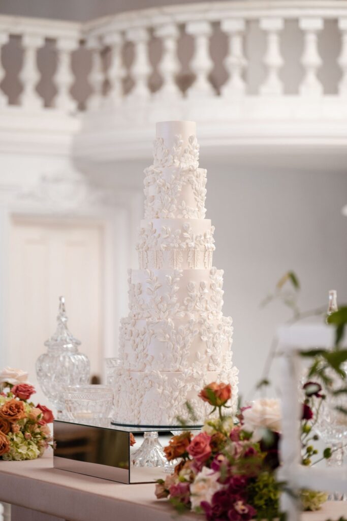 Six-tier white wedding cake covered in sugar leaf and flower appliqués, with roses in the foreground