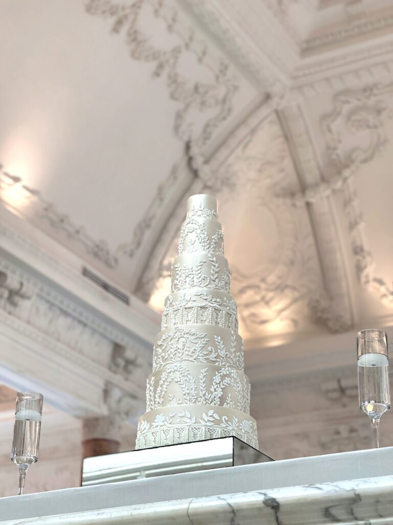 Seven-tier pearl wedding cake with piped floral lace beneath an ornate plaster ceiling