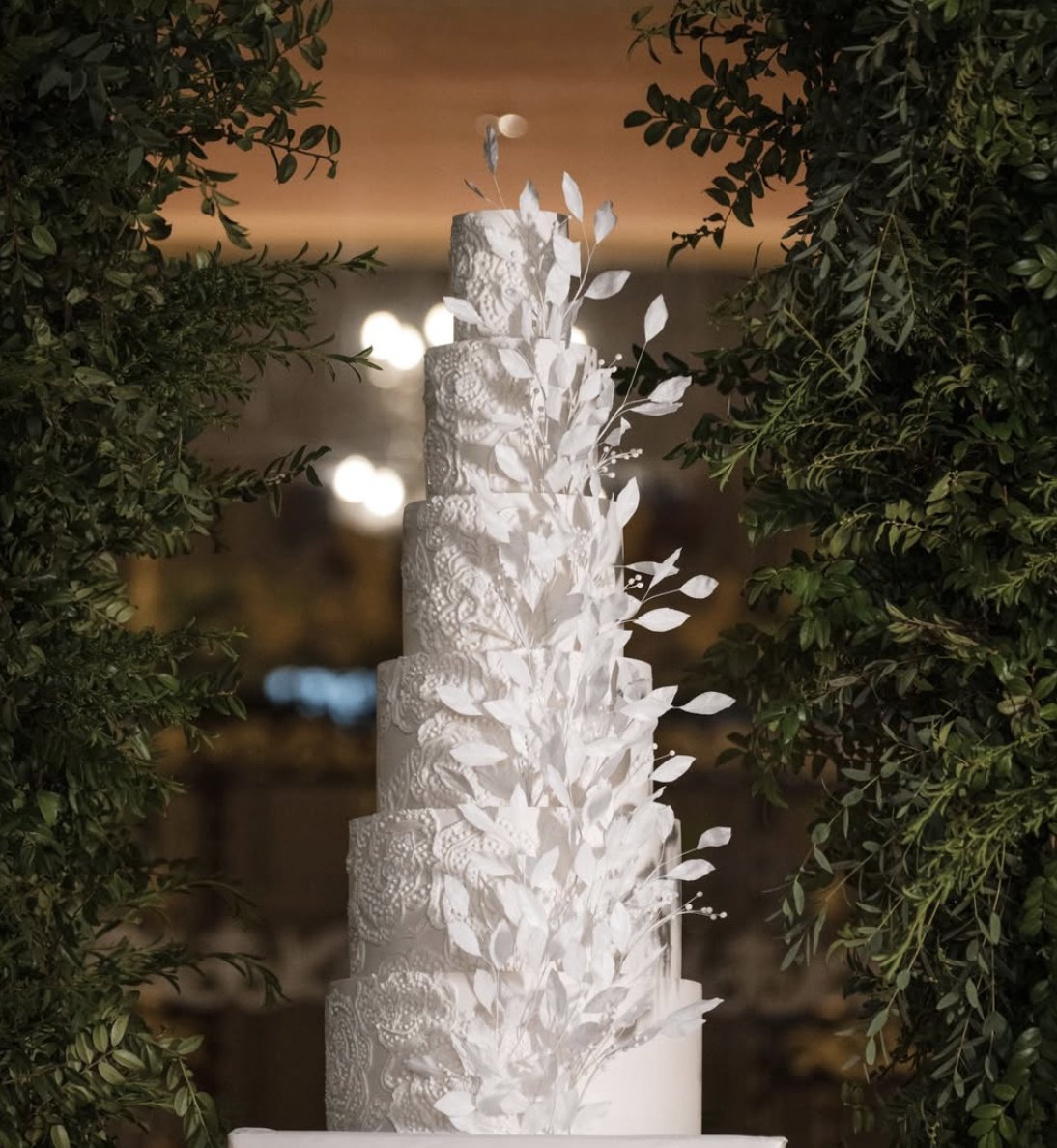Tall white wedding cake covered in cascading sugar leaves, framed by greenery
