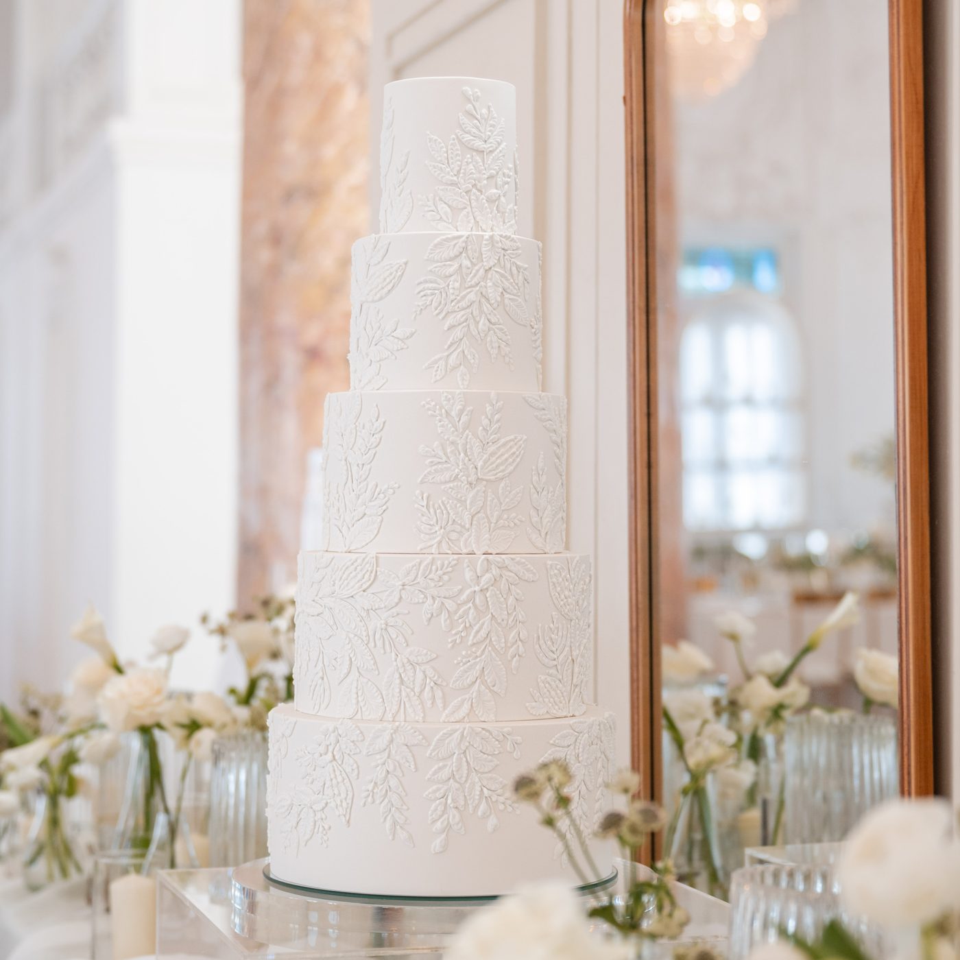 Five-tier white wedding cake with raised leaf piping beside a wooden-framed mirror